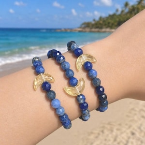 May include: Three beaded bracelets with blue and yellow accents. Each bracelet features a mix of blue gemstone beads and a yellow crescent moon charm. The bracelets are displayed on a person's wrist, with a beach and ocean in the background.