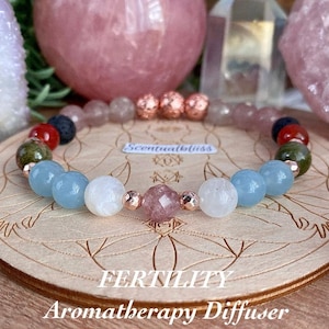 May include: A gemstone bead bracelet featuring pink, blue, white, red, and green beads. The bracelet is presented on a wooden surface. The words "FERTILITY Aromatherapy Diffuser" and the brand name "Scentualbliss" are visible.