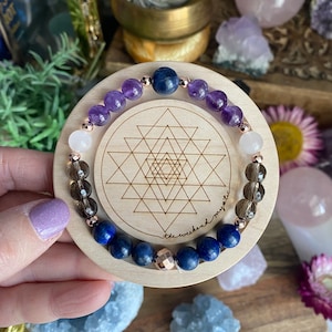 May include: A gemstone bracelet with amethyst, rose quartz, smoky quartz, and lapis lazuli beads. The bracelet has rose gold-coloured accents and is displayed on a wooden coaster with a geometric pattern. The coaster has the text "the weekend mystic".