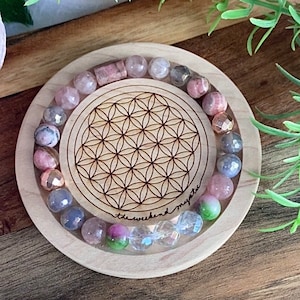 May include: A wooden tray with a flower of life design engraved on it. A bracelet made of pink, white, and gray stones is displayed on the tray. The text "the weekend mystic" is written on the tray.