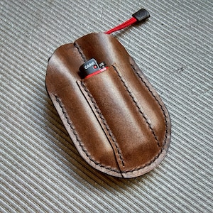 May include: A brown leather case with a red pull tab and a single pocket for a lighter. The lighter is visible in the pocket and has the word "Cricket" printed on it.