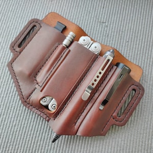 Puede incluir: Una funda de cuero marrón para multiherramientas con una multiherramienta, un bolígrafo y una linterna. La funda tiene múltiples bolsillos y ranuras, con clips metálicos para asegurar los artículos. El cuero está cosido con hilo negro.