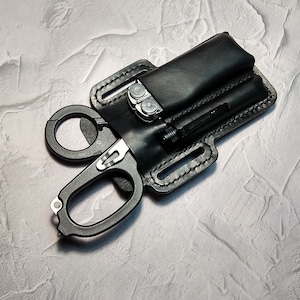 Könnte beinhalten: Eine schwarze Lederscheide mit einem Multitool, einer Taschenlampe und einer schwarzen Schere.