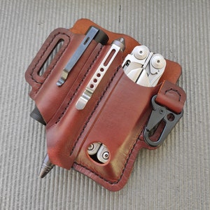 Könnte beinhalten: Ein braunes Lederetui für Multitools mit einem Clip und einer Tasche für einen Stift. Das Etui hat eine Klappe, die mit einem Druckknopf geschlossen wird.