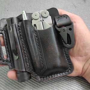 May include: A black leather multi-tool and flashlight holder with a clip. The holder has three pockets, one for a flashlight, one for a multi-tool, and one for a pen. The holder is made of distressed leather and has a brown patina.