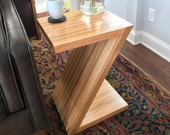 May include: A wooden end table with a Z-shaped design. The table has a light wood finish and a small shelf. A white coffee mug sits on a round coaster on the top shelf.