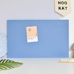 May include: A rectangular blue magnetic memo board featuring a peach-coloured Pantone 14-1119 Winter Wheat paper swatch attached with two small grey star-shaped magnets. The board has a smooth, solid blue surface and is positioned horizontally.