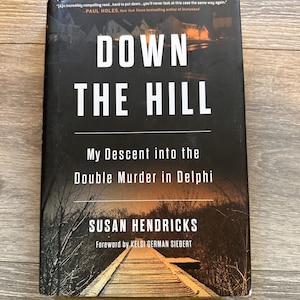 Puede incluir: Un libro titulado "DOWN THE HILL" de Susan Hendricks. La cubierta oscura presenta texto blanco y una ilustración de un puente. El subtítulo es "My Descent into the Double Murder in Delphi".