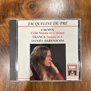 Puede incluir: Una caja de CD con Jacqueline Du Pré. La portada incluye el texto "CHOPIN Cello Sonata in G minor, FRANCK Sonata in A, DANIEL BARENBOIM." La caja de CD tiene un lomo negro y una foto de una mujer con cabello largo.