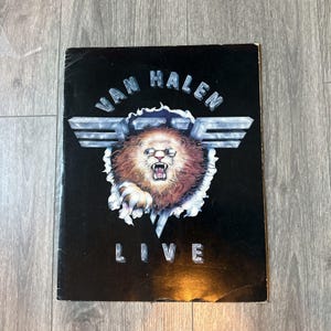 Può includere: Una copertina di un album nera con la scritta "VAN HALEN LIVE" in argento. La copertina presenta un leone ruggente con gli occhiali, che sfonda un effetto carta strappata, con ali argentate sopra il leone.