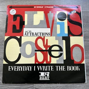May include: A vintage vinyl record album cover for "Elvis Costello and the Attractions." The cover features bold typography in red, black, and gray, with the title "Everyday I Write the Book." The record is a 12-inch single with 3 tracks.