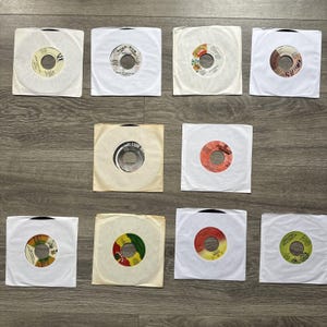 May include: Twelve vinyl records in white paper sleeves are arranged on a wooden surface. The record labels feature various colours and designs, including red, yellow, and green. The records are in their original sleeves.