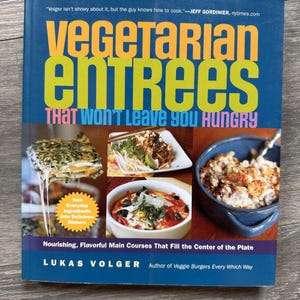 May include: A cookbook titled "Vegetarian Entrees That Won't Leave You Hungry" by Lukas Volger. The cover is blue with large yellow and orange text. It features images of various vegetarian dishes, promoting nourishing and flavorful main courses.