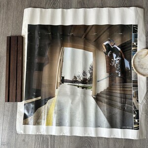 May include: A large poster featuring a skateboarder mid-air, performing a trick on a staircase. The poster has a white border and is creased. A set of dark wooden slats and a small, round object are next to the poster.