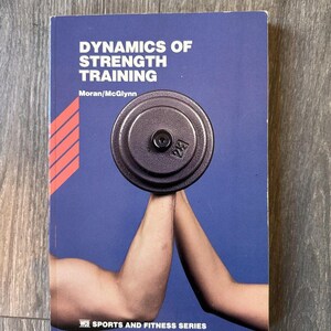 May include: A book titled "Dynamics of Strength Training" with a blue cover. The cover features a barbell weight held by two arms, with the weight's number "25" visible. The book is part of the "WCB Sports and Fitness Series."