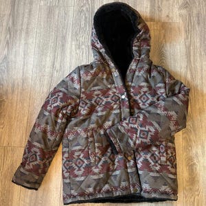 Könnte beinhalten: Kapuzenjacke mit geometrischem Muster in Braun-, Grau- und Burgundertönen. Die Jacke hat ein schwarzes Kunstfellfutter und einen Knopfverschluss. Die Jacke hat zwei Taschen und ist für kaltes Wetter konzipiert.