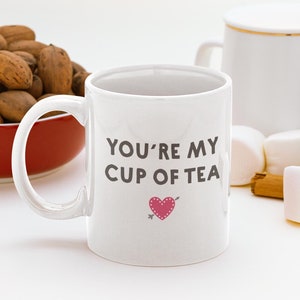 May include: White ceramic mug with the text "YOU'RE MY CUP OF TEA" in grey, featuring a pink heart with an arrow. The mug is next to a red bowl of nuts, marshmallows, and cinnamon sticks.