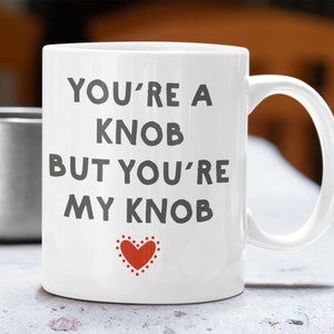 May include: White ceramic mug with the text "YOU'RE A KNOB BUT YOU'RE MY KNOB" in dark grey lettering. A small red heart with white dots is below the text. A silver mug is in the background.