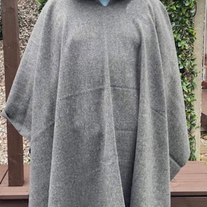 May include: A grey hooded poncho made of a thick, textured fabric. The garment features a pointed hood and a straight hemline finished with a thin, contrasting red trim along the bottom edge. The poncho drapes loosely over the shoulders.