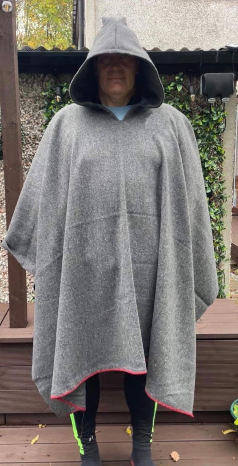 May include: A grey hooded poncho made of a thick, textured fabric. The garment features a pointed hood and a straight hemline finished with a thin, contrasting red trim along the bottom edge. The poncho drapes loosely over the shoulders.