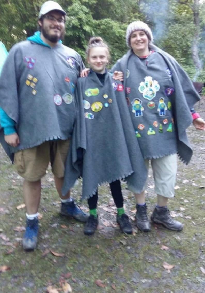 May include: Three grey felt ponchos featuring various colourful embroidered patches and pins attached to the front. Each poncho has a jagged, unfinished hemline and is worn over casual clothing including shorts, leggings, and hiking boots.