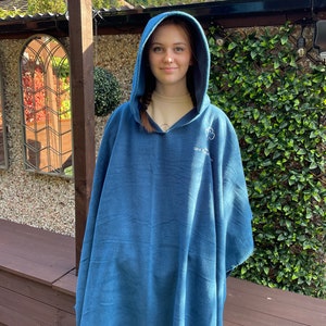 May include: A blue hooded fleece poncho featuring a white embroidered clover emblem and text on the upper chest area. The garment is made of a soft, solid-coloured fabric and includes a hood that drapes over the shoulders and down the torso.