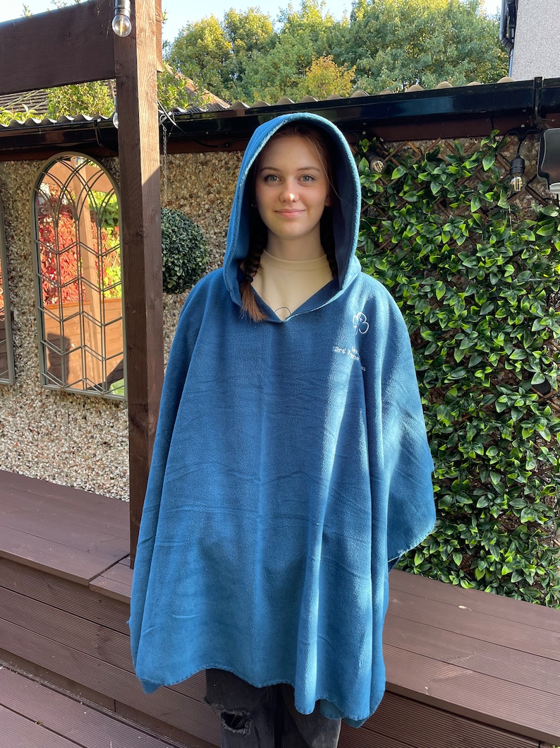 May include: A blue hooded fleece poncho featuring a white embroidered clover emblem and text on the upper chest area. The garment is made of a soft, solid-coloured fabric and includes a hood that drapes over the shoulders and down the torso.