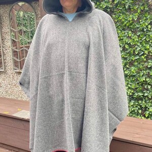 May include: A grey hooded poncho made of a thick, textured fabric. The garment features a pointed hood and a straight hemline finished with a thin red trim along the bottom edge. The poncho drapes loosely over the shoulders and torso.