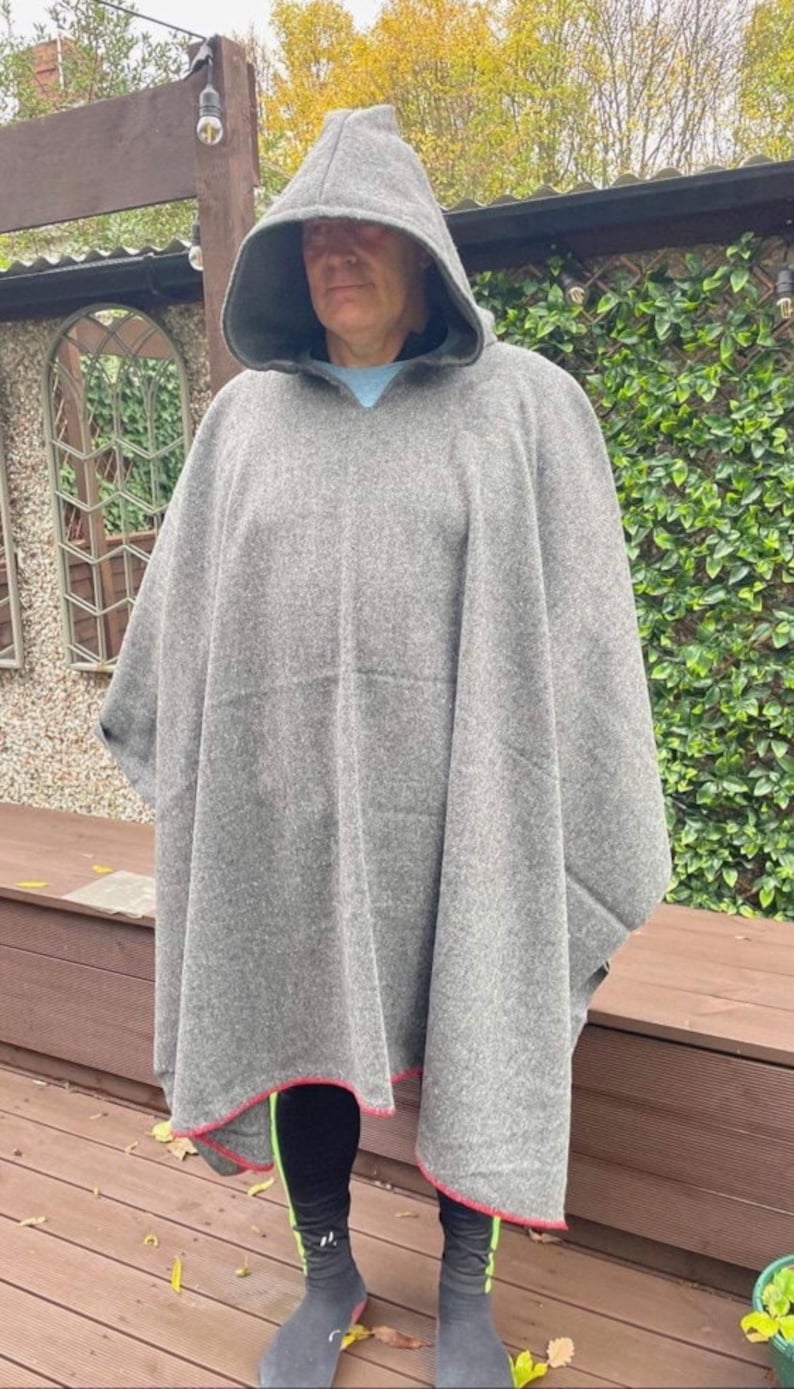 May include: A grey hooded poncho made of a thick, textured fabric. The garment features a pointed hood and a straight hemline finished with a thin red trim along the bottom edge. The poncho drapes loosely over the shoulders and torso.