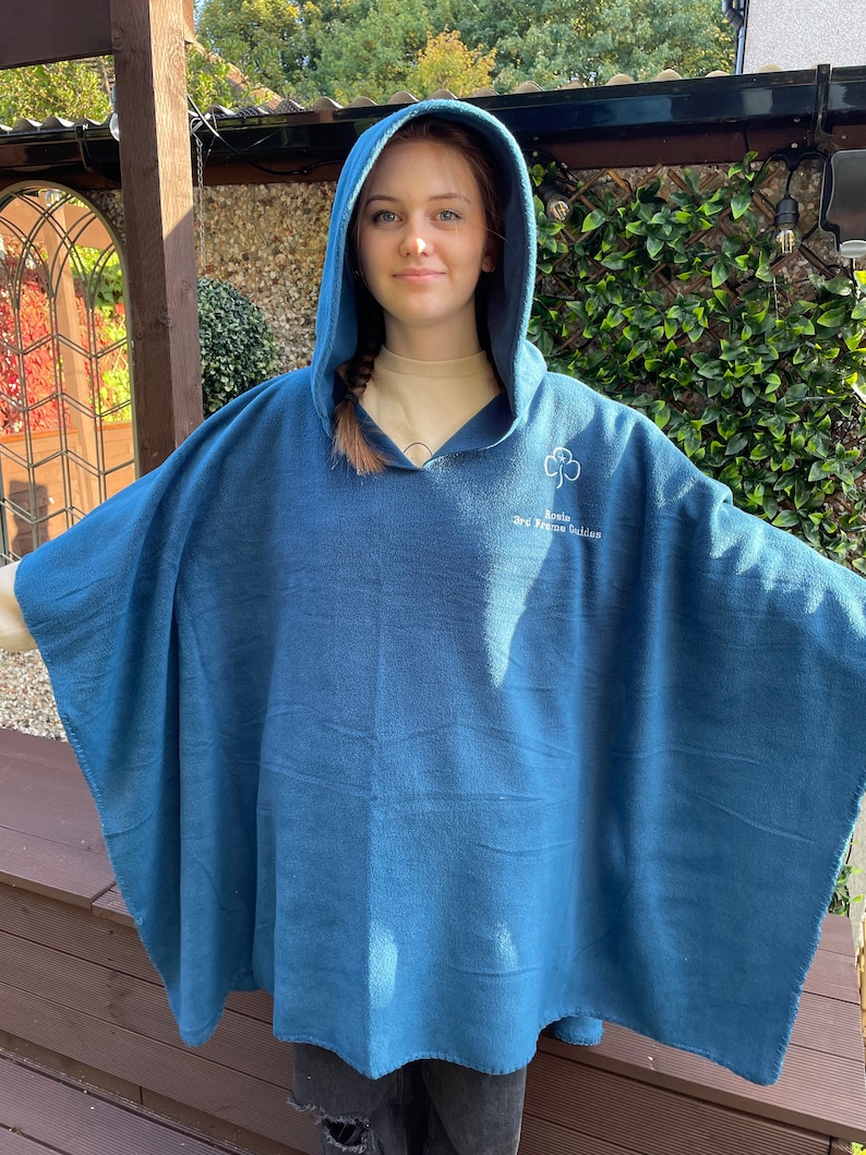 May include: A blue fleece hooded poncho featuring an embroidered white trefoil logo and the text "Rosin 3rd Frame Guides" on the upper chest. The garment has a wide, loose-fitting silhouette with an attached hood and long, open sides.