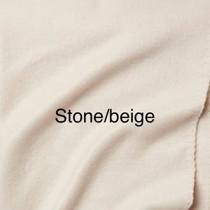 May include: A soft, solid-coloured fabric blanket in a light stone or beige hue. The material features a smooth, slightly fuzzy texture with gentle folds and ripples across the surface. The text Stone/beige is printed in black.