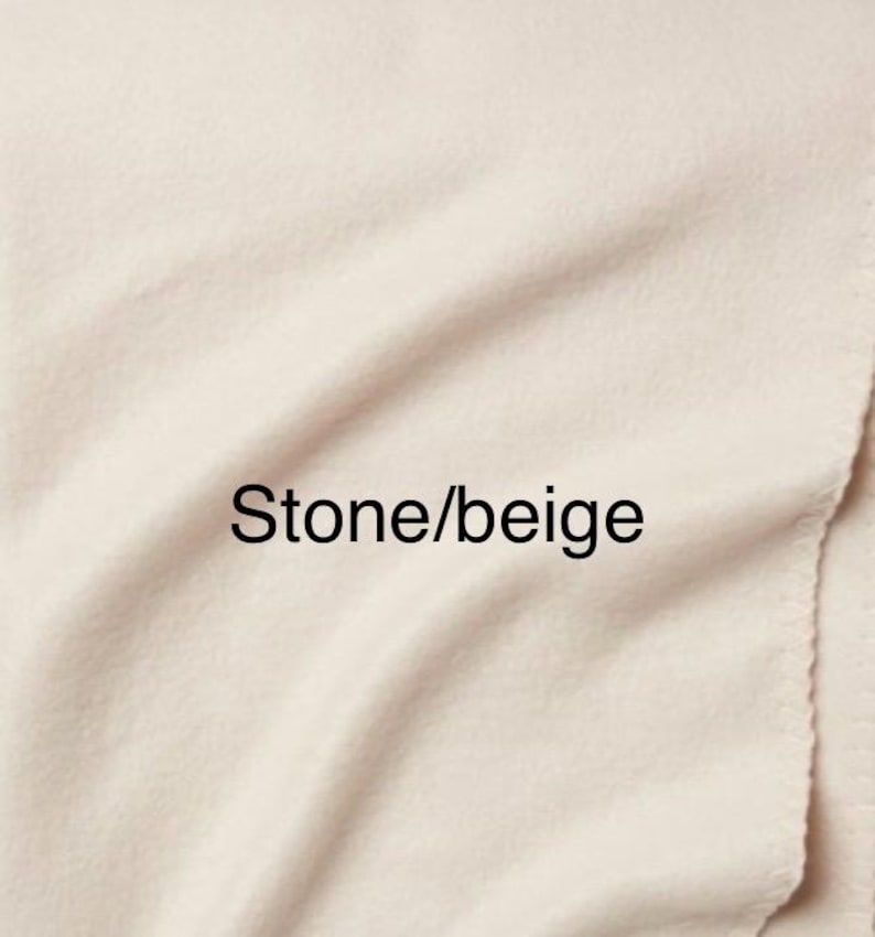 May include: A soft, solid-coloured fabric blanket in a light stone or beige hue. The material features a smooth, slightly fuzzy texture with gentle folds and ripples across the surface. The text Stone/beige is printed in black.