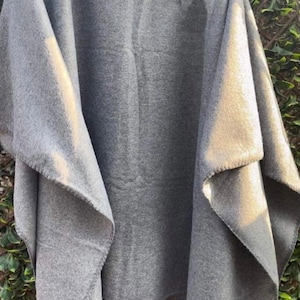 May include: A solid grey poncho made of a soft, felted fabric. The garment features a wide, draped silhouette with an open front design and a simple collar, hanging from a wooden hanger to display its loose, flowing structure.