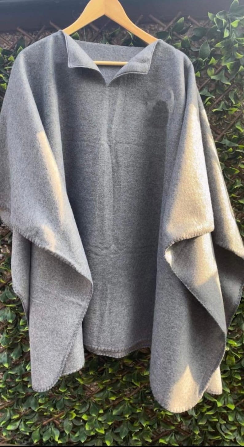 May include: A solid grey poncho made of a soft, felted fabric. The garment features a wide, draped silhouette with an open front design and a simple collar, hanging from a wooden hanger to display its loose, flowing structure.