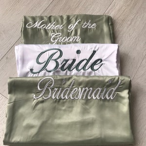 May include: Three folded satin robes, each embroidered with text. The top robe is sage green and says "Mother of the Groom" in white. The middle robe is white with "Bride" in teal. The bottom robe is sage green with "Bridesmaid" in white.