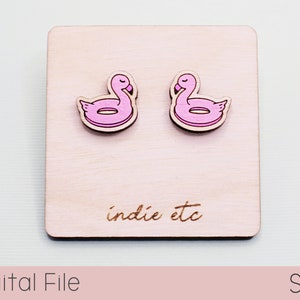 May include: A pair of wooden earrings featuring a pink flamingo pool float design. The earrings are displayed on a wooden surface with the text "indie etc" engraved on it.