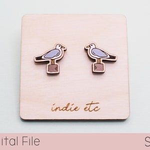 May include: A pair of wooden earrings featuring a seagull perched on a post. The earrings are light blue and brown with a white outline. The earrings are displayed on a wooden surface with the text "indie etc" engraved on the surface.