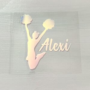 May include: A clear, square sticker with a cheerleader silhouette in a jumping pose, holding pom-poms. The name "Alexi" is written in cursive. The design has a holographic, iridescent finish, with a light pink and yellow colour gradient.