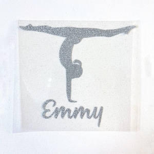 Pode incluir: Silhueta prateada com glitter de uma ginasta em posição de parada de mãos. O nome "Emmy" está escrito em itálico abaixo. O design está em um fundo transparente e ligeiramente texturizado.