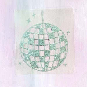 Puede incluir: Un diseño de bola de discoteca verde brillante con un patrón de cuadros y cuatro acentos de estrellas. La imagen está sobre un fondo suave y texturizado con una combinación de colores rosa claro y blanco. La bola de discoteca tiene un lazo para colgar.