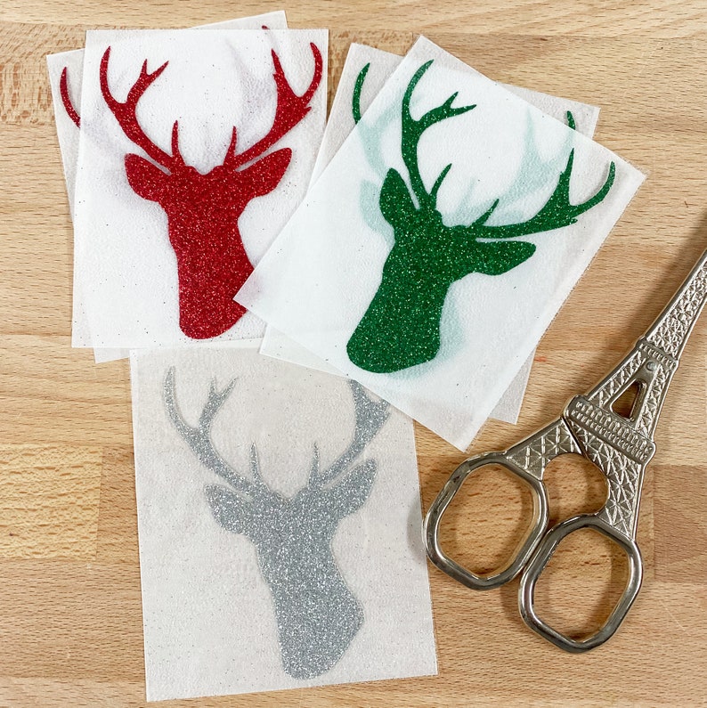 Op de afbeelding: Verschillende glitterende hertenkop-stickers in rood, groen en zilver worden getoond op een licht houten oppervlak. Een decoratieve schaar met een Eiffeltoren-ontwerp is zichtbaar. De stickers zitten op een witte achtergrond.