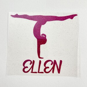 May include: A pink glitter decal featuring a silhouette of a gymnast performing a handstand with the name ELLEN printed in a bold, cursive font underneath the figure. The design is presented on a clear, adhesive backing material.