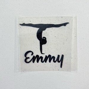 May include: A black glittery iron-on transfer decal featuring a silhouette of a gymnast performing a handstand. Below the gymnast, the name Emmy is written in a black glittery cursive script. The design is set on a clear adhesive backing.