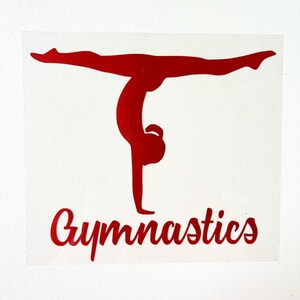 May include: A red vinyl decal featuring a silhouette of a gymnast performing a handstand with legs in a wide split. Below the figure, the word Gymnastics is written in a bold, cursive red font. The design is presented on a clear backing sheet.