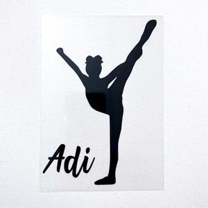 以下が含まれることがあります： 片腕を上げてハイキックをするチアリーダーのシルエットが描かれた黒いビニールステッカー。チアリーダーの図の左側には、透明な台紙の上に黒い筆記体フォントで「Adi」という名前が印刷されています。