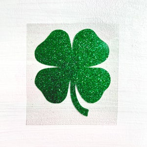May include: A green glitter four-leaf clover design on a clear square. The clover is a vibrant green color and sparkles. The background is a white, textured surface.
