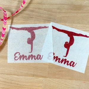 May include: Two rectangular iron-on transfers featuring a glittery pink and a glittery red silhouette of a gymnast performing a handstand split. Below each gymnast figure, the name Emma is printed in a cursive script font in matching glittery colors.