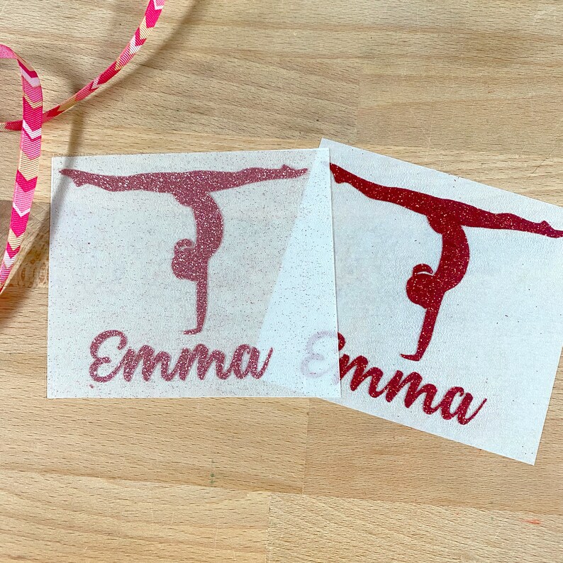 May include: Two rectangular iron-on transfers featuring a glittery pink and a glittery red silhouette of a gymnast performing a handstand split. Below each gymnast figure, the name Emma is printed in a cursive script font in matching glittery colors.