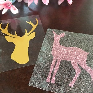 Op de afbeelding: Twee vinyl stickers met silhouetten van herten. E&eacute;n is een gouden bokkenkop, de andere een roze glitterhert. Ze liggen op een donkere ondergrond, met roze bloemen op de achtergrond.