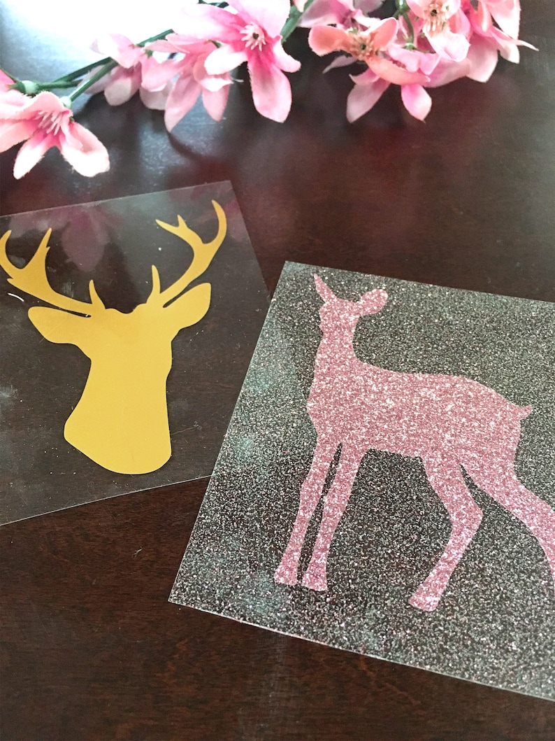 Op de afbeelding: Twee vinyl stickers met silhouetten van herten. E&eacute;n is een gouden bokkenkop, de andere een roze glitterhert. Ze liggen op een donkere ondergrond, met roze bloemen op de achtergrond.